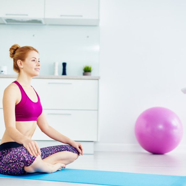 Portrait of fit red haired woman doing yoga exercises at home on floor: sitting with legs crossed in lotus position on mat and smiling