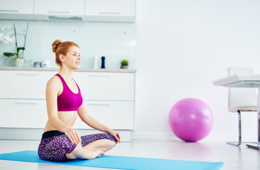 Portrait of fit red haired woman doing yoga exercises at home on floor: sitting with legs crossed in lotus position on mat and smiling